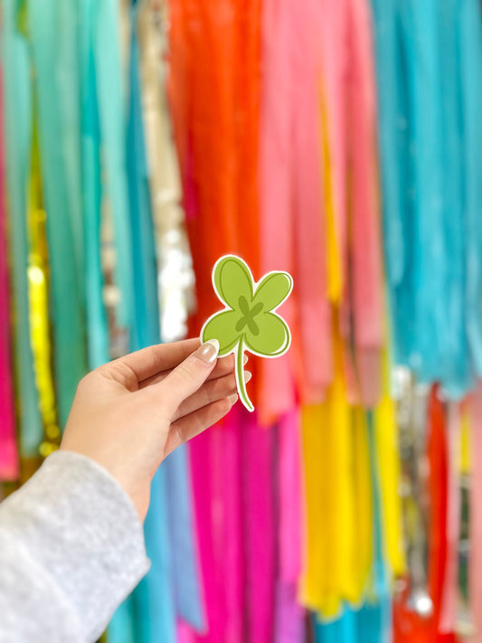 A hand holding a green 4 leaf clover sticker in front of a colorful backdrop with streamers.
