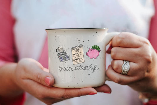 A hand holding a white ceramic mug with an illustration of accounting-themed items like a calculator, paperwork, and a pink fruit, along with the text '#ACCOUNTANTLIFE'.