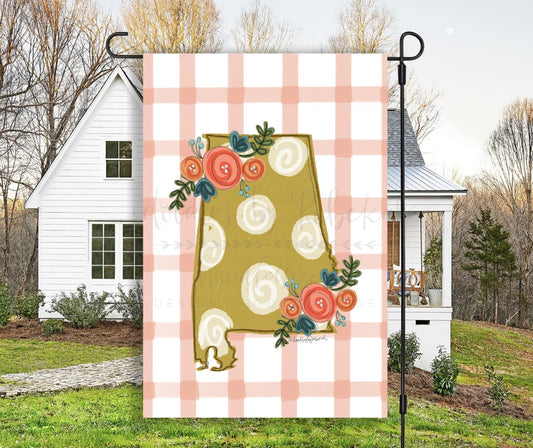 A garden flag with a plaid pattern and floral dot design, featuring the state of Alabama outline with floral accents, displayed in an outdoor setting.