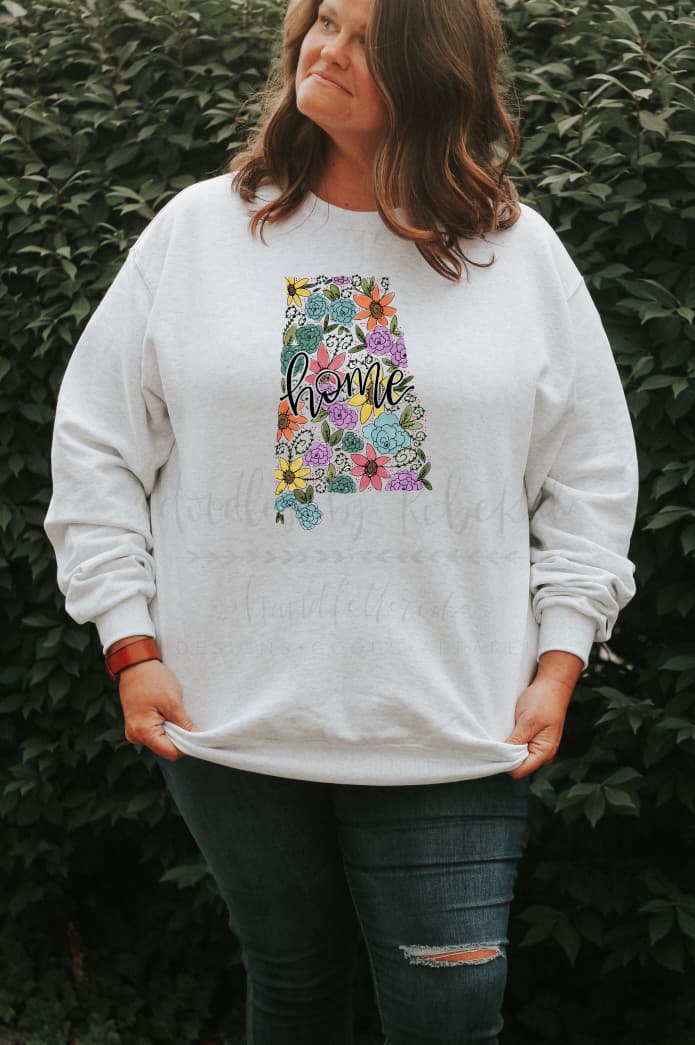 a woman standing wearing a white sweatshirt with a floral design in the shape of Alabama 