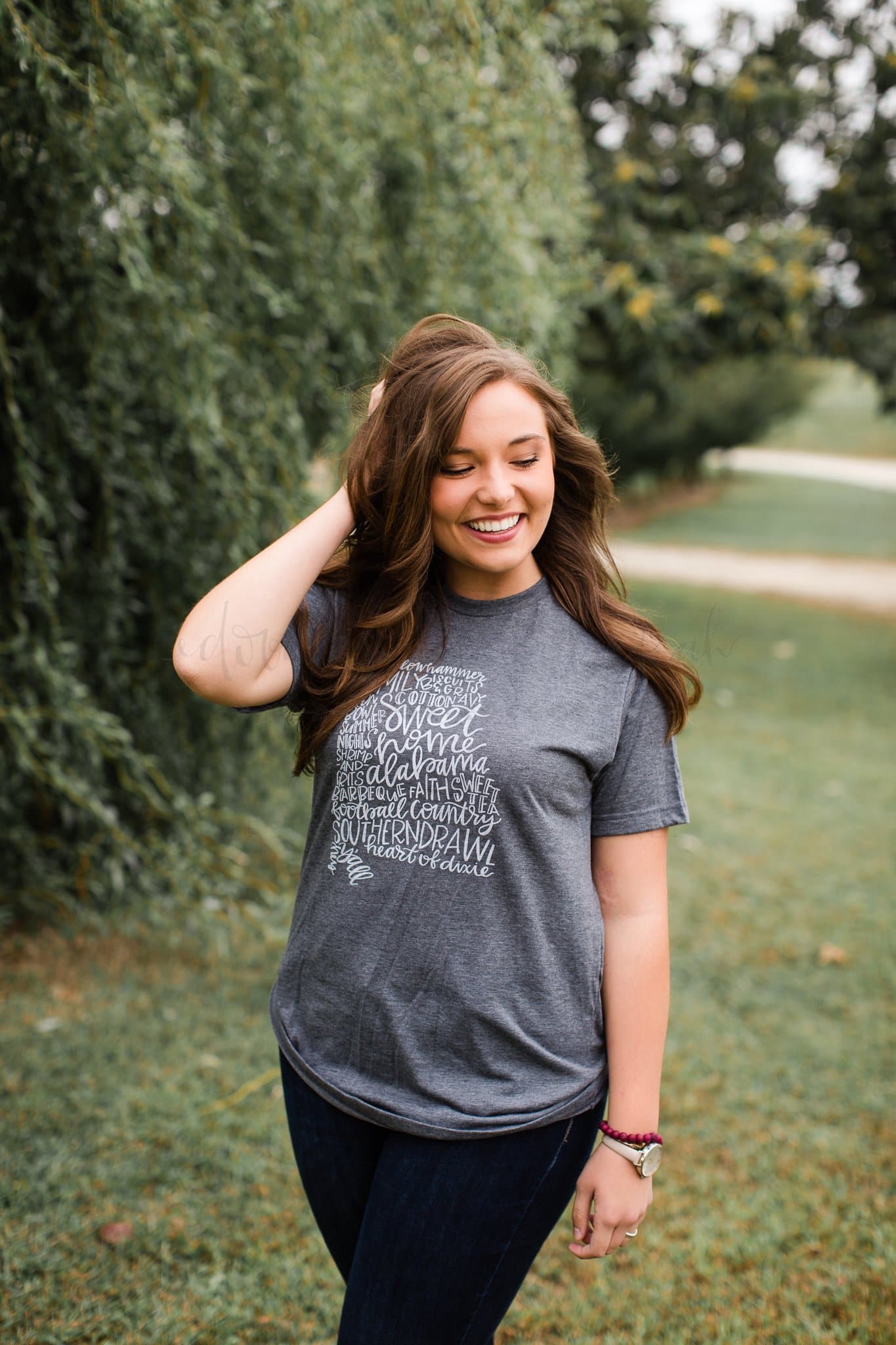 woman standing outside in a grey shirt with cursive writing on it in the shape of Alabama