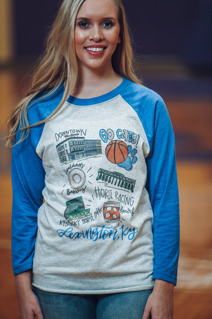 A woman wearing a blue and white long-sleeved baseball t-shirt with hand-drawn Kentucky-themed graphics, standing indoors.