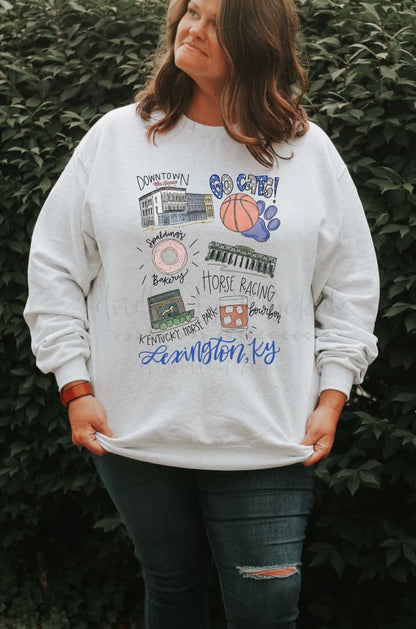 A woman standing outside wearing a white sweatshirt with hand drawn graphics representing the city of Lexington, Kentucky