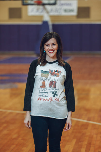 A person standing in an indoor location wearing a white raglan t-shirt with hand-drawn lettering and designs.