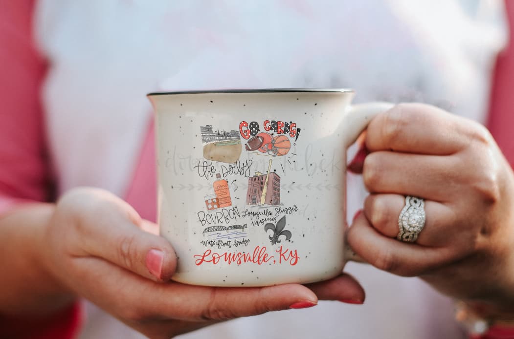 A person holding a beige ceramic mug with text and decorative elements related to Louisville, KY.