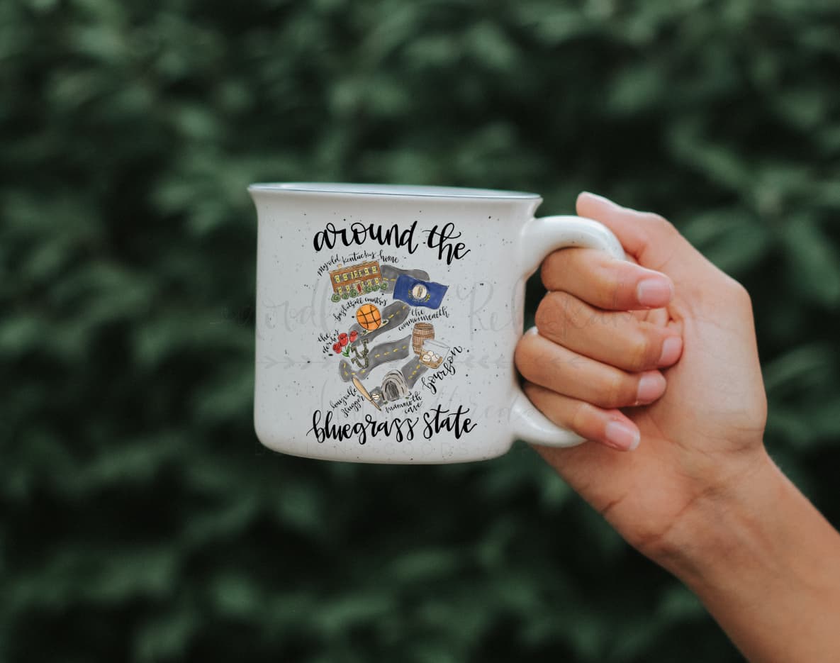 A hand holding a white ceramic mug with text and a graphic related to Kentucky.