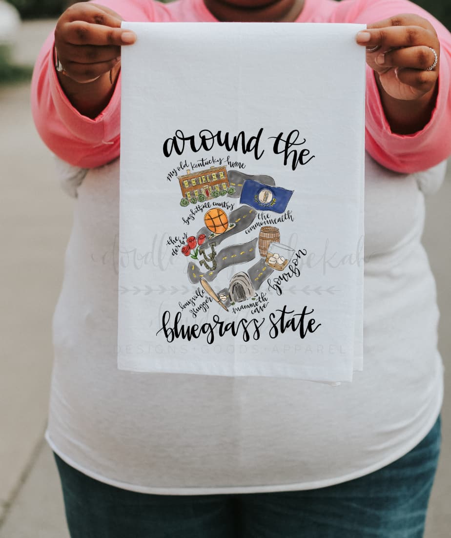 A person holding a white polyester tea towel with 'Around the Bluegrass State' printed in black text and various illustrations related to Kentucky.