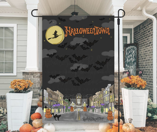 A garden flag with a Halloween theme, featuring a black background with orange and white text, depicting a witch, bats, and a small town at night. Displayed in a yard.
