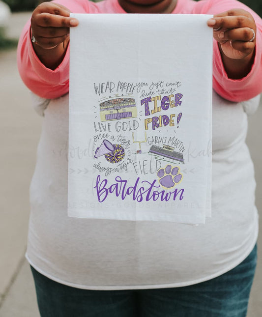 A person holding up a white polyester tea towel with purple and pink text and graphics related to Bardstown High School pride.