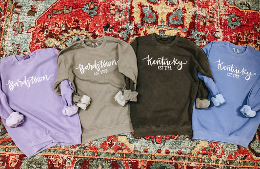 A collection of four sweatshirts in gray, purple, blue, and brown colors, each with the text 'BARDSTOWN' or 'KENTUCKY' printed on the front.