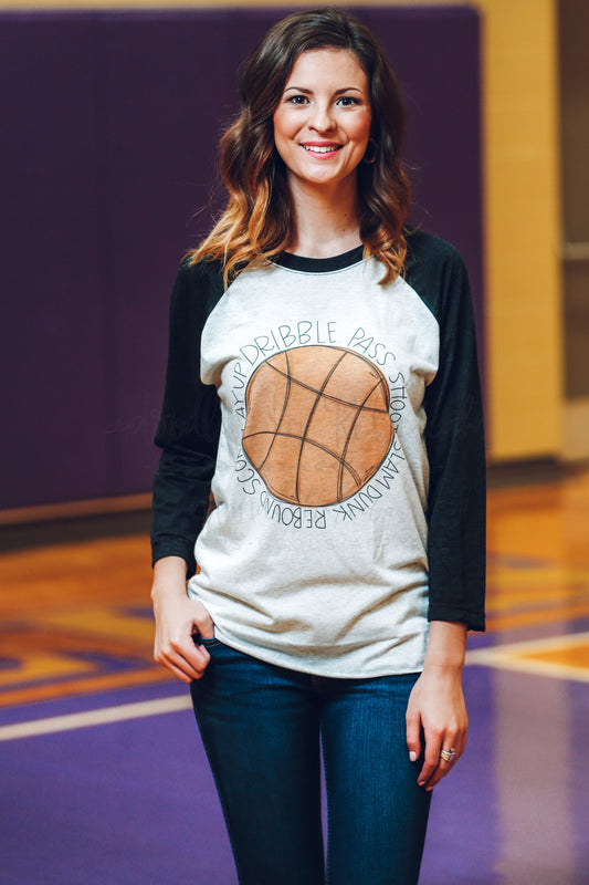 A woman smiling and wearing a beige t-shirt with a basketball graphic and the phrases 'Dribble. Pass.