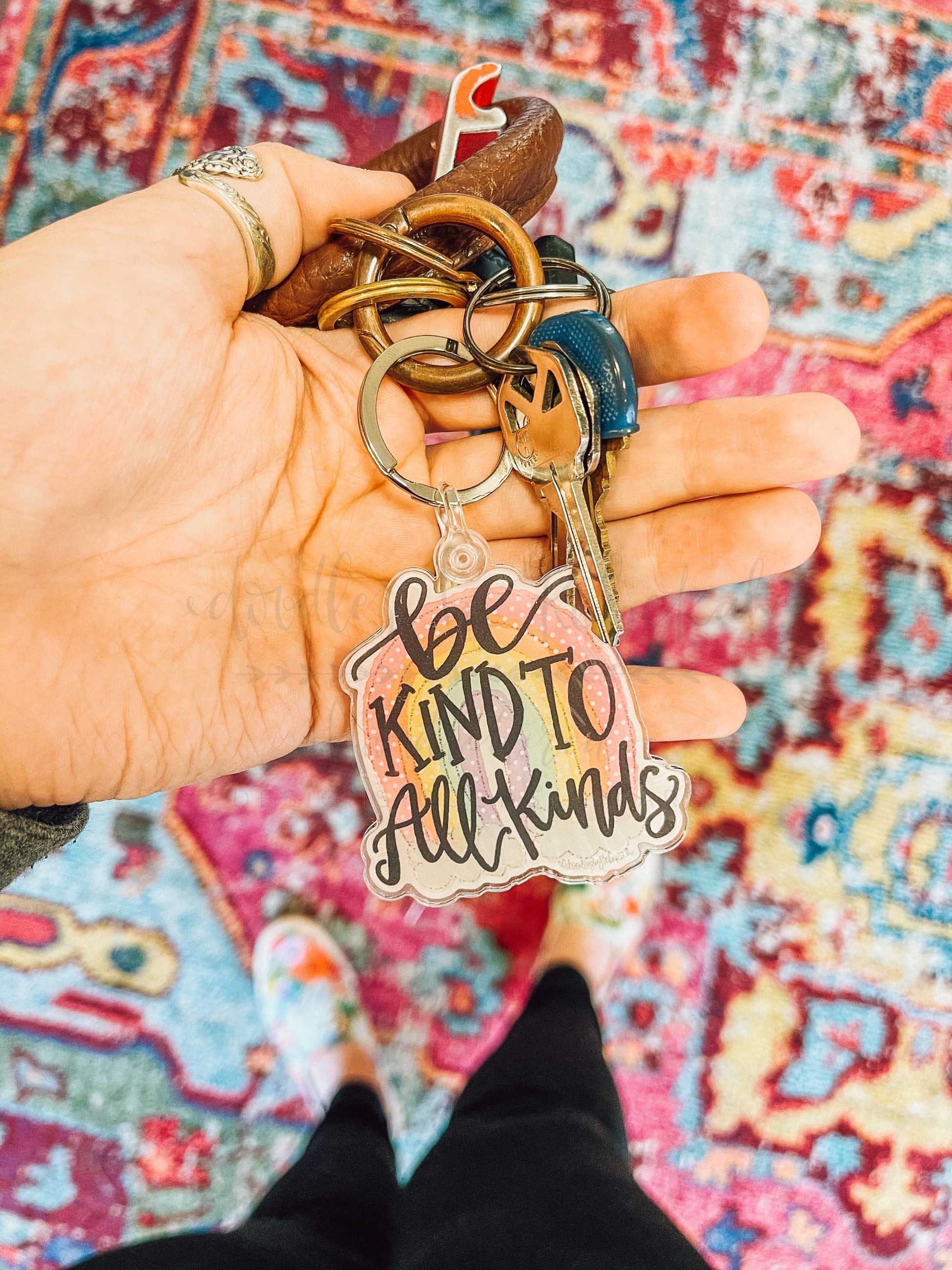 An acrylic keychain with a metal ring, featuring the phrase 'BE KIND TO ALL KINDS' in a cursive script.