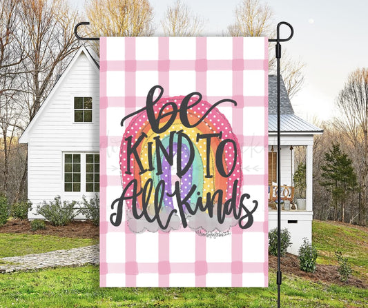 A garden flag with a pink plaid background and the text 'BE KIND TO ALL KINDS' in the center in a black scripted font.