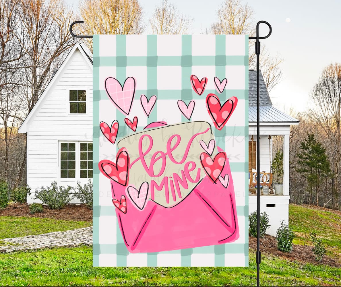 A garden flag with a design of a pink envelope with hearts and the phrase 'Be Mine' on it, displayed in an outdoor setting.