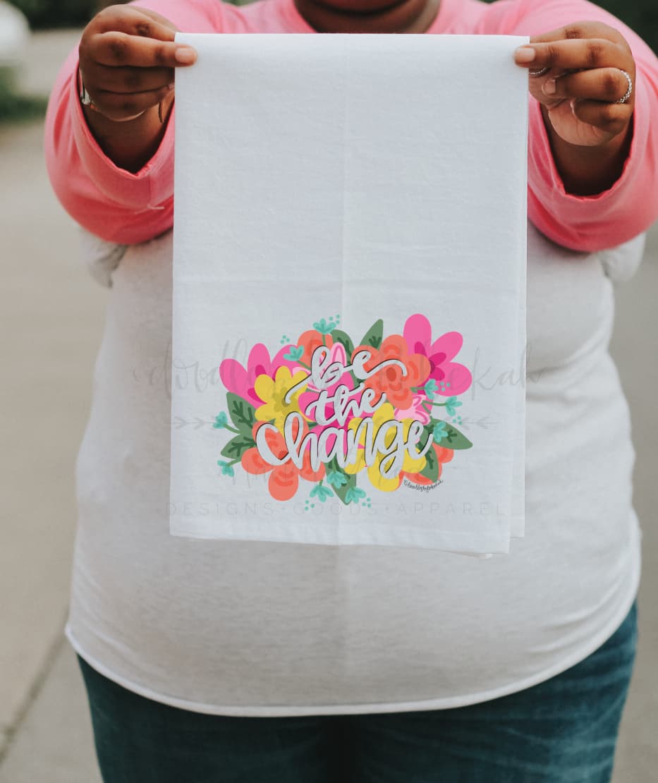 A person holding a white polyester tea towel with a colorful floral design and the text 'BE THE CHANGE' on it.