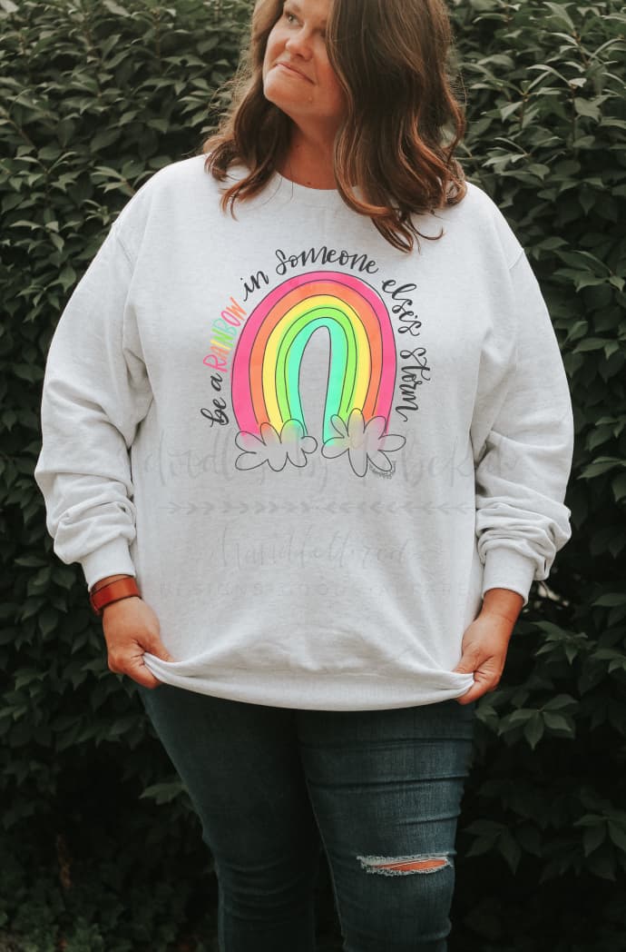 A person wearing a white crew neck t-shirt with a colorful text design that reads 'be a rainbow in someone else's storm'.