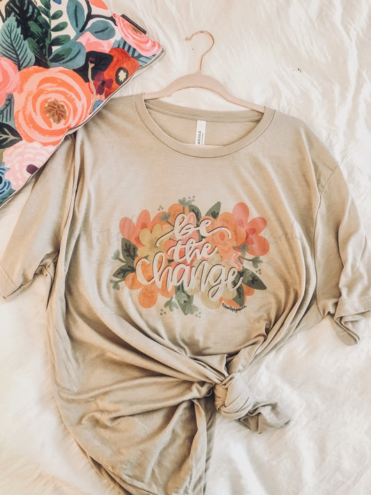 A beige colored t-shirt with hand-drawn lettering and floral designs, displayed on a flat surface.
