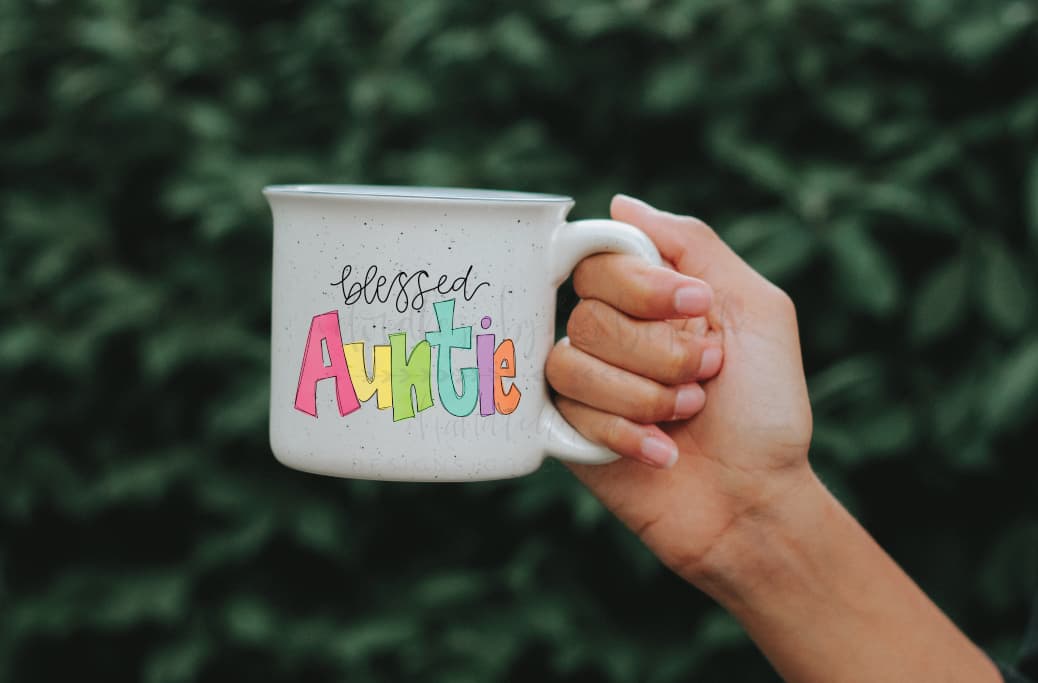 A hand holding a white ceramic mug with a colorful 'Blessed Auntie' text design on it.