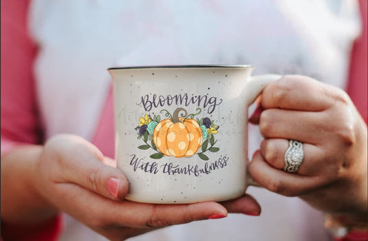 A person holding a white ceramic mug with a floral and pumpkin design and the text 'Blooming with Thankfulness'.