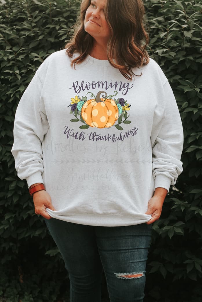 A woman wearing a white sweatshirt with a pumpkin on it that reads "Blooming in Thankfulness"