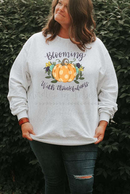 A woman wearing a white sweatshirt with a pumpkin on it that reads "Blooming in Thankfulness"