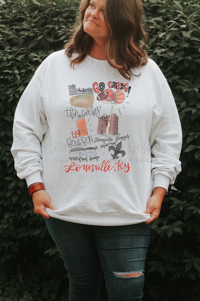 A person standing in an indoor location wearing a white sweatshirt with hand-drawn lettering and designs representing Louisville, Kentucky