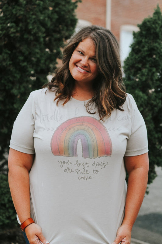 A smiling person wearing a beige t-shirt with hand-drawn lettering and a rainbow design.