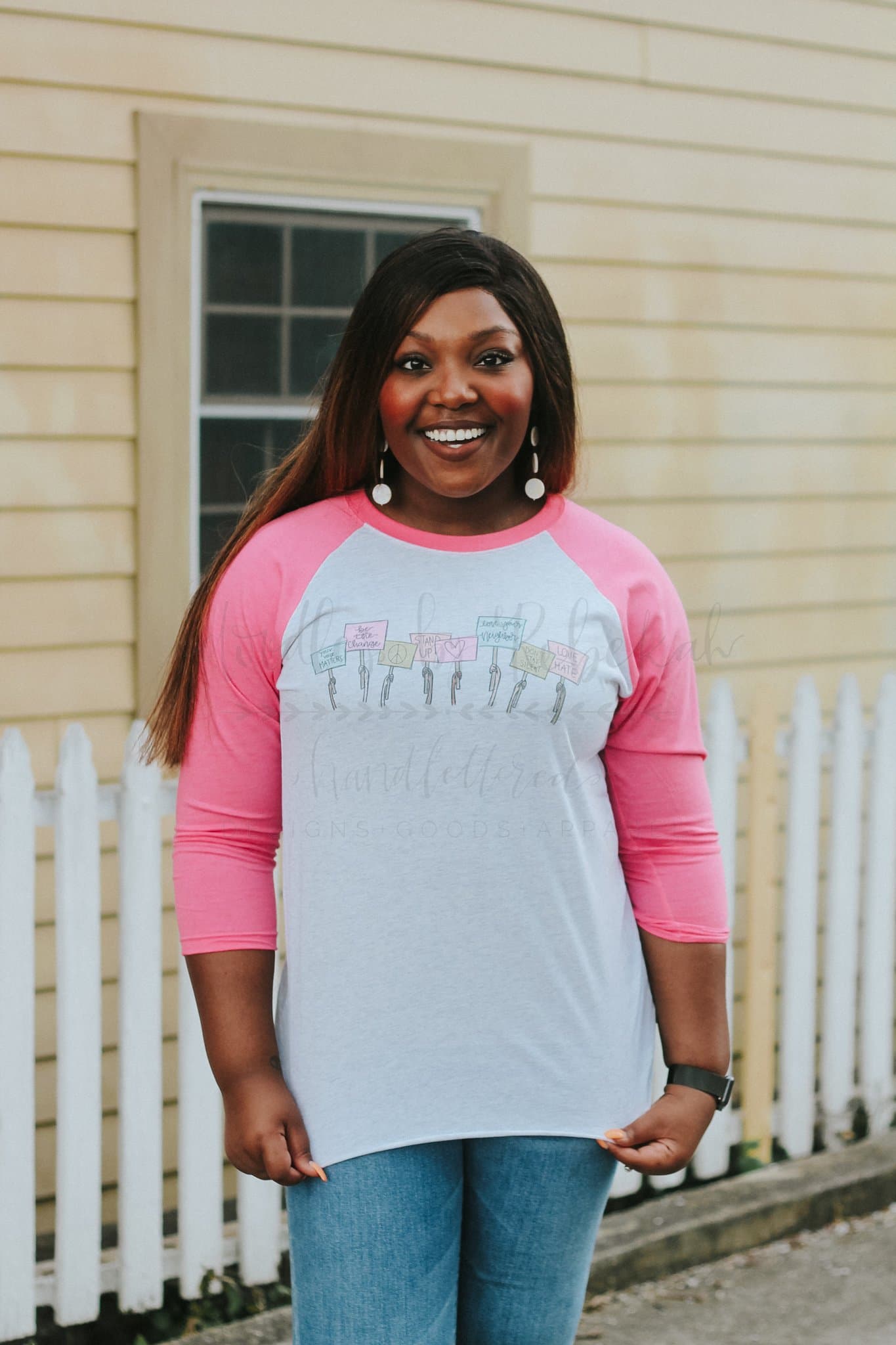 A person wearing a long-sleeved pink and gray t-shirt with hand-drawn lettering and designs, standing in front of a white picket fence.