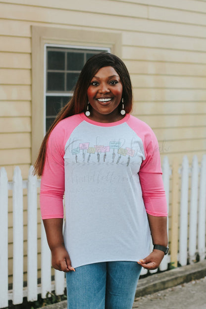 A person wearing a long-sleeved pink and gray t-shirt with hand-drawn lettering and designs, standing in front of a white picket fence.