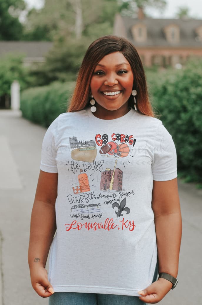 A person standing in an outdoor location wearing a white  t-shirt with hand-drawn lettering and designs representing Louisville, Kentucky